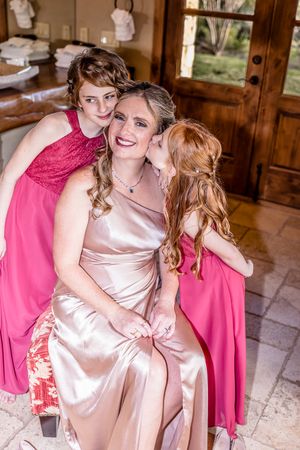 Three people share a tender moment wearing formal dresses in pink and champagne satin at a special event.