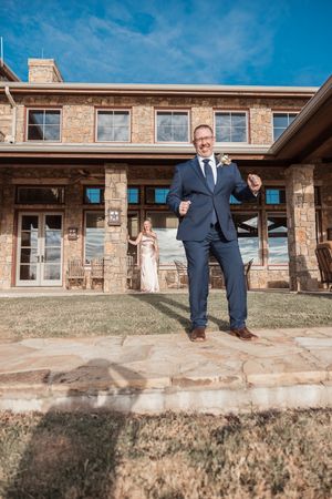 A person in a navy suit poses confidently outside a rustic stone building on a sunny day with blue skies.