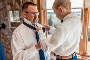 Two men in white shirts getting ready for formal event with one adjusting the other's blue necktie.