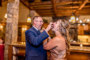 A couple shares a playful moment while feeding each other cake in a rustic venue with warm lighting.