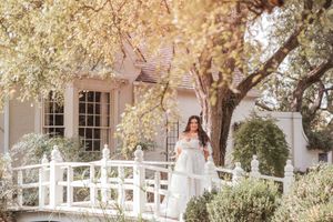 A figure in a white dress stands on an arched wooden bridge in front of a cottage surrounded by blooming trees.