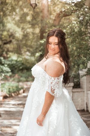 A bride in a white lace off-shoulder wedding gown poses along a sunlit garden path with dappled lighting.
