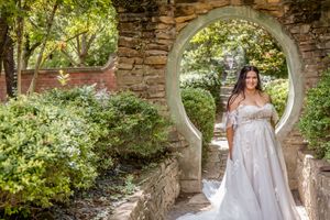 A person in a white wedding dress poses under a circular stone archway surrounded by lush green foliage on a sunny day.