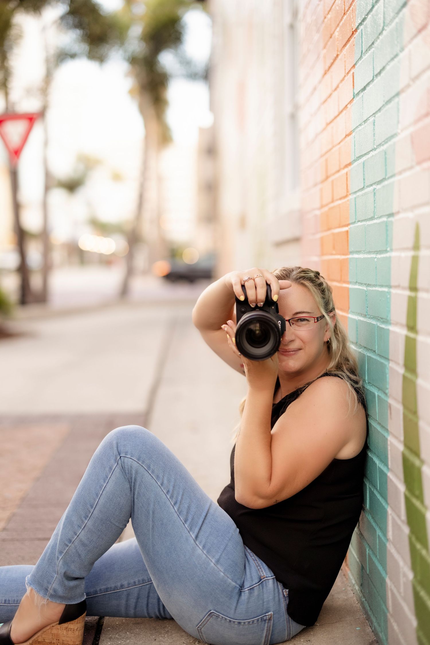 Female photographer sitting against colorful tiled wall holding DSLR camera on urban sidewalk.