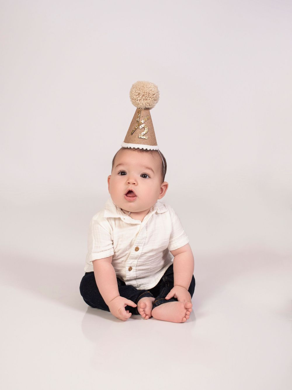 Baby wearing white onesie with a crown accessory on head sitting against light background.