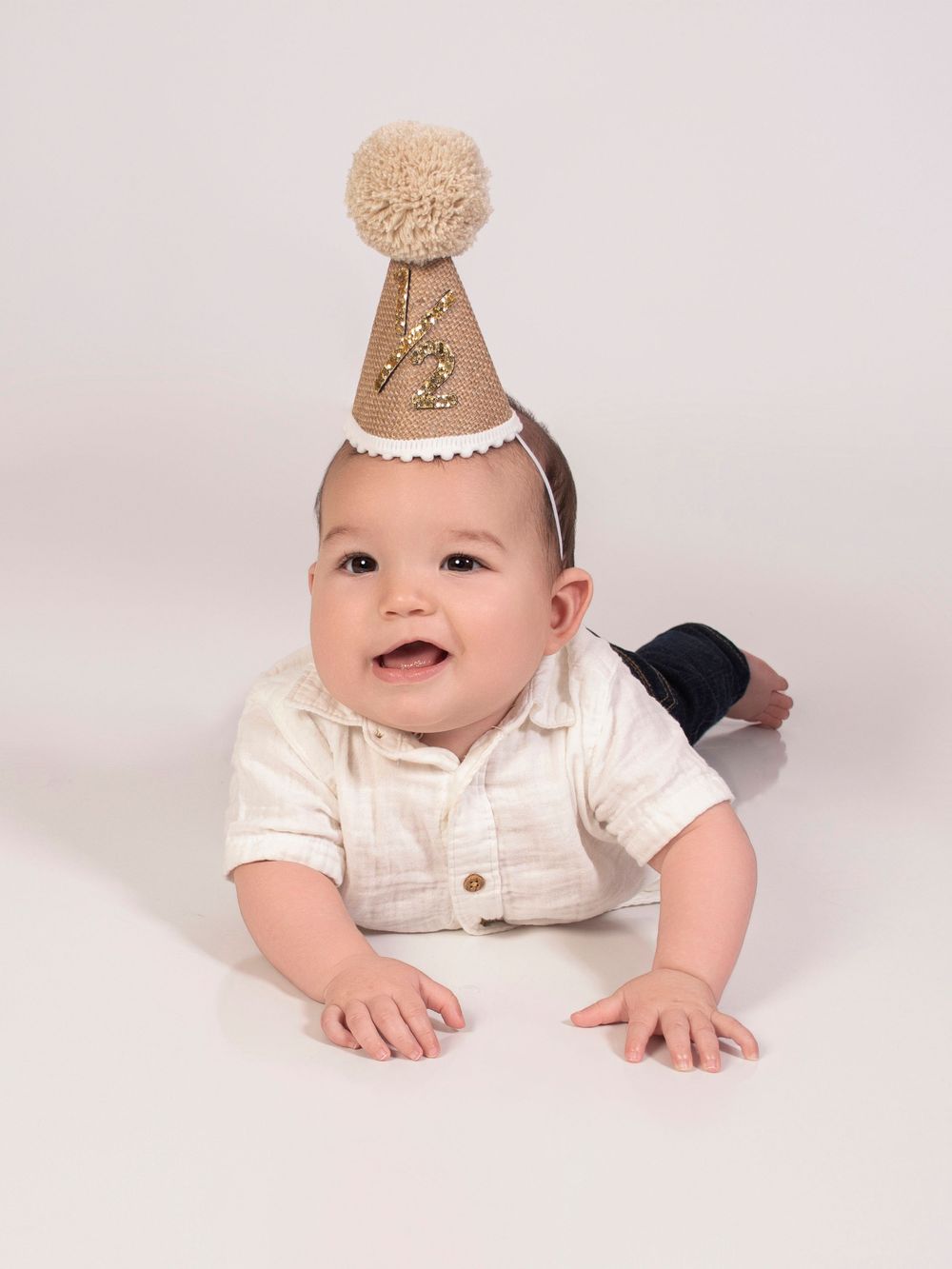 A baby wearing a light-colored outfit and paper crown lies on their stomach on a white background in a series of poses.