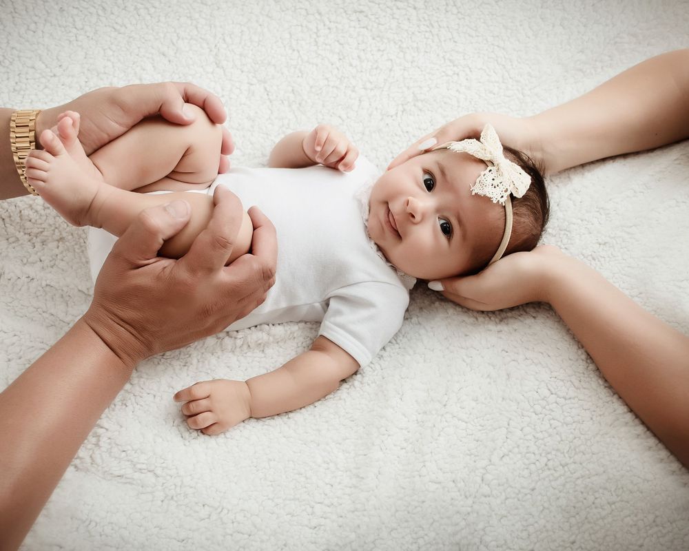 Hands gently hold a peaceful baby wearing white clothing and lying on soft textured fabric.