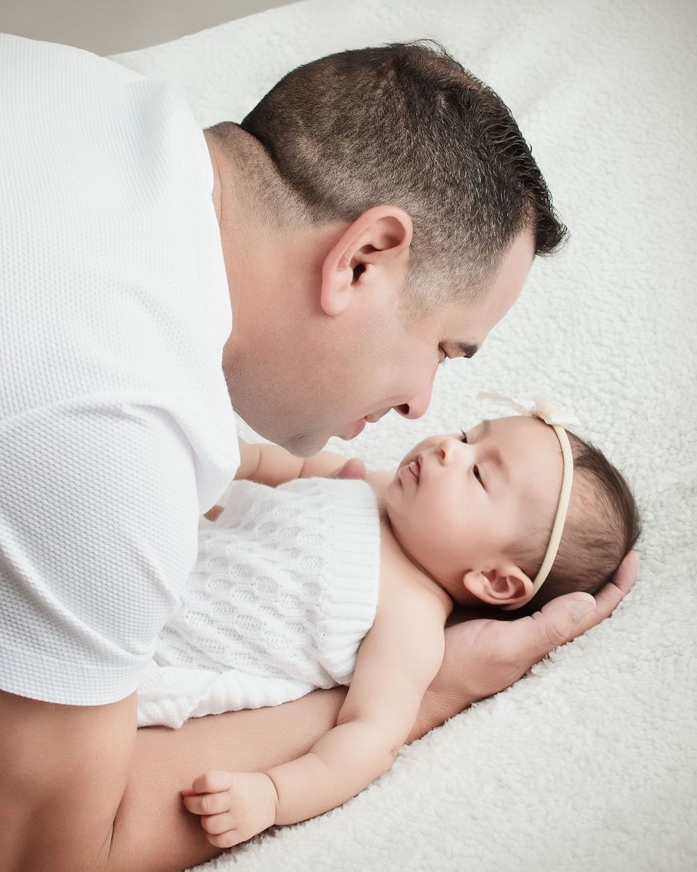 A tender moment of parent sharing a gentle kiss with sleeping baby wearing white headband on textured white bedding.