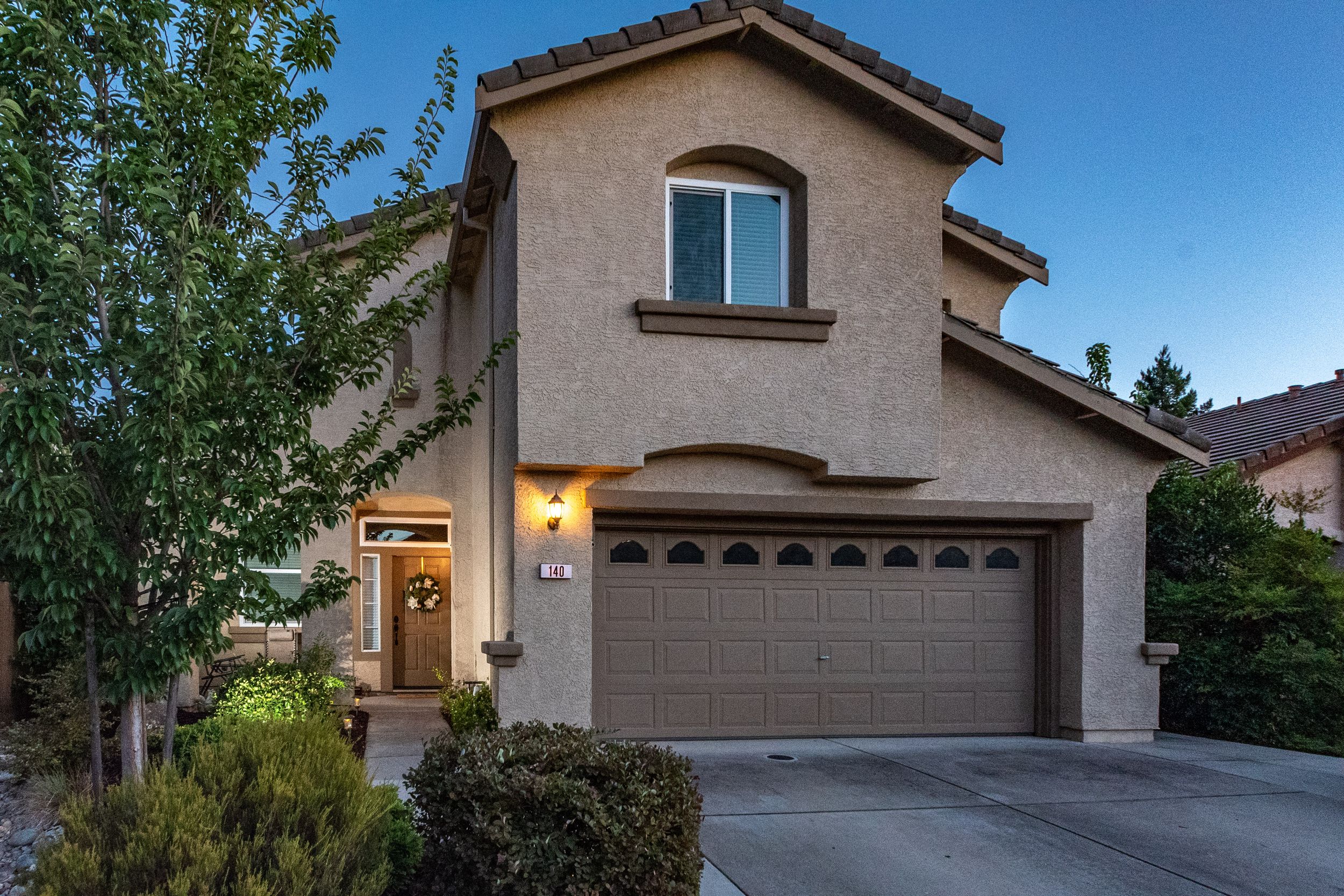 Two-story stucco home with tan garage door, arched windows, and lush trees at dusk in a residential neighborhood.