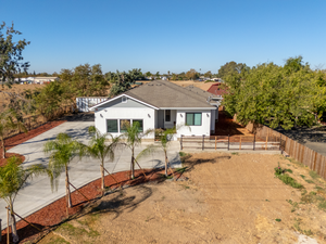 Aerial view of a single-story white house with gray roof, concrete driveway, young palm trees, and a fenced yard under a clear blue sky.
