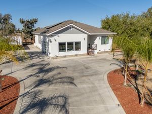 Single-story white house with solar panels, large windows, and a wide concrete driveway surrounded by palm trees and red mulch landscaping.