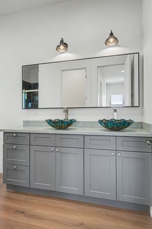 Gray double vanity with two blue-green glass vessel sinks, a large frameless mirror, and black wall sconces above a wood-look floor.