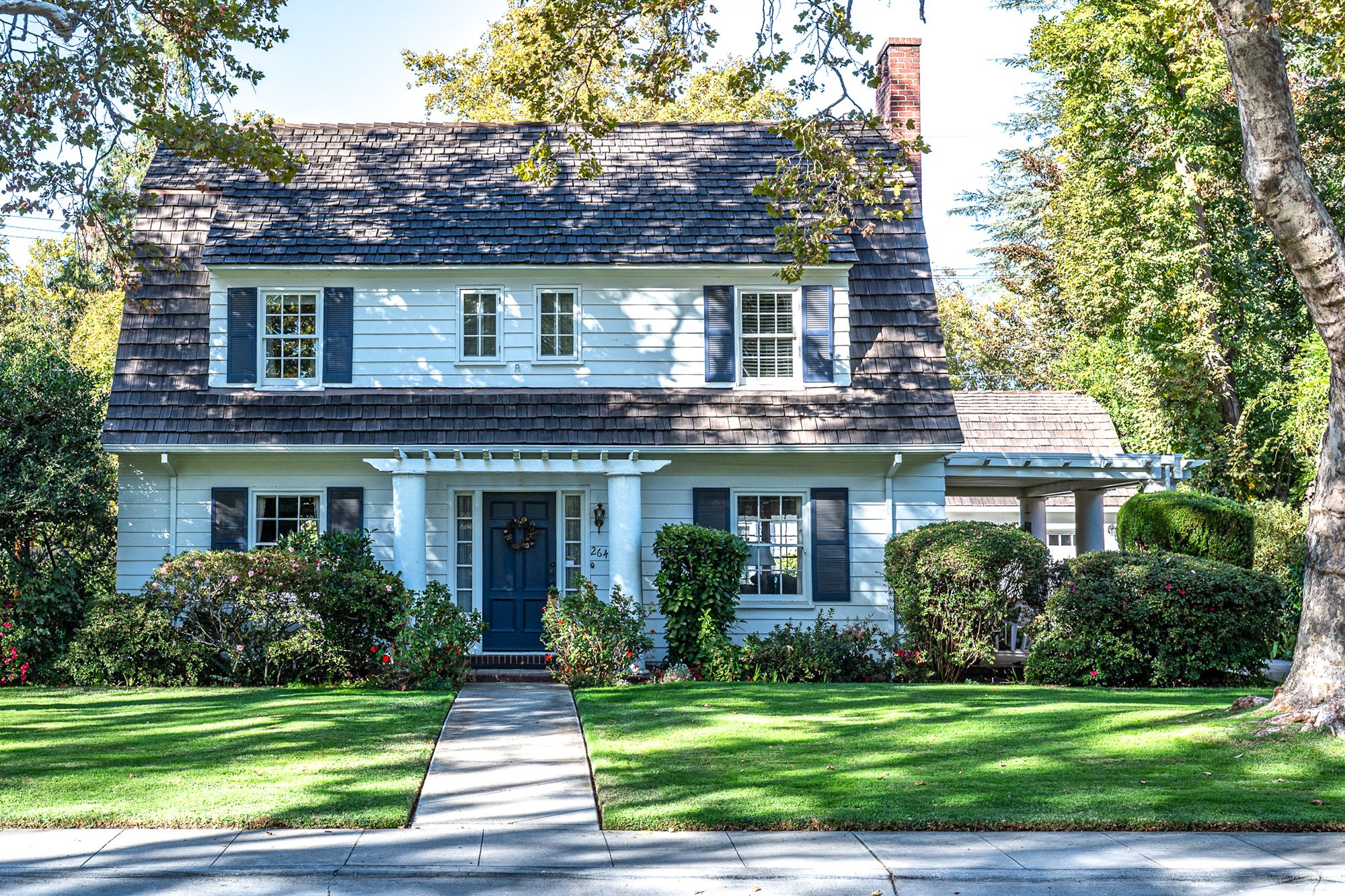 Charming light blue two-story home with dark shutters, a shingled roof, a front portico and a landscaped green lawn beside a walkway.