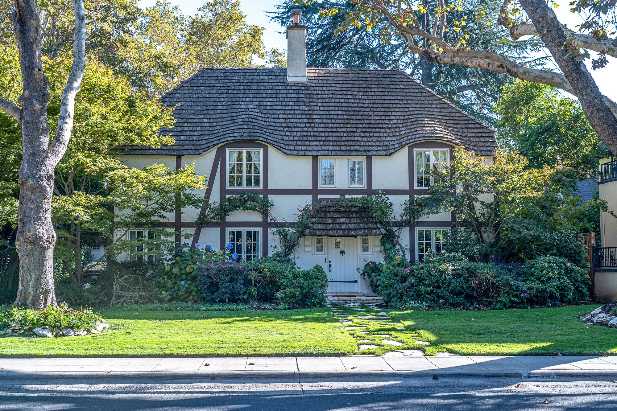 Tudor-style two-story home with half-timbered facade, shingled roof, and brick chimney, framed by mature trees and a manicured front lawn.