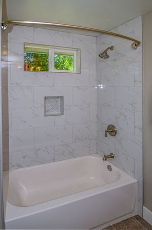 White marble tiled shower and bathtub combo with curved brass rod, showerhead, and small window in elegant bathroom.