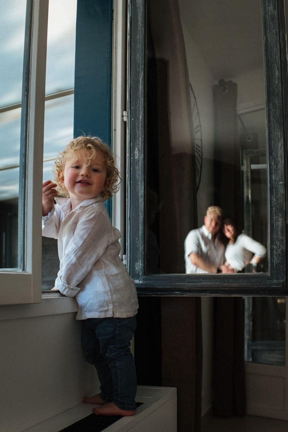 enfant jouant à la fenetre sous le regard de ses parents en seance photo de famille a domicile a Lille