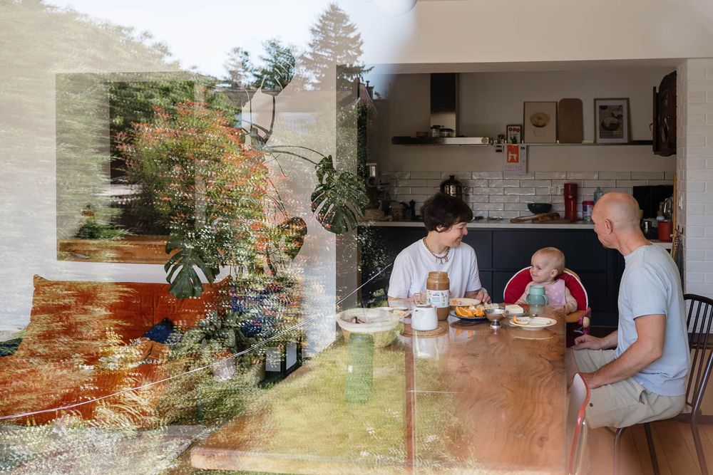 Belle photo avec un reflet du jardin : parents prenant leur petit dejeuner avec leur bebe de 1 an, par une photographe de famille a Lille.