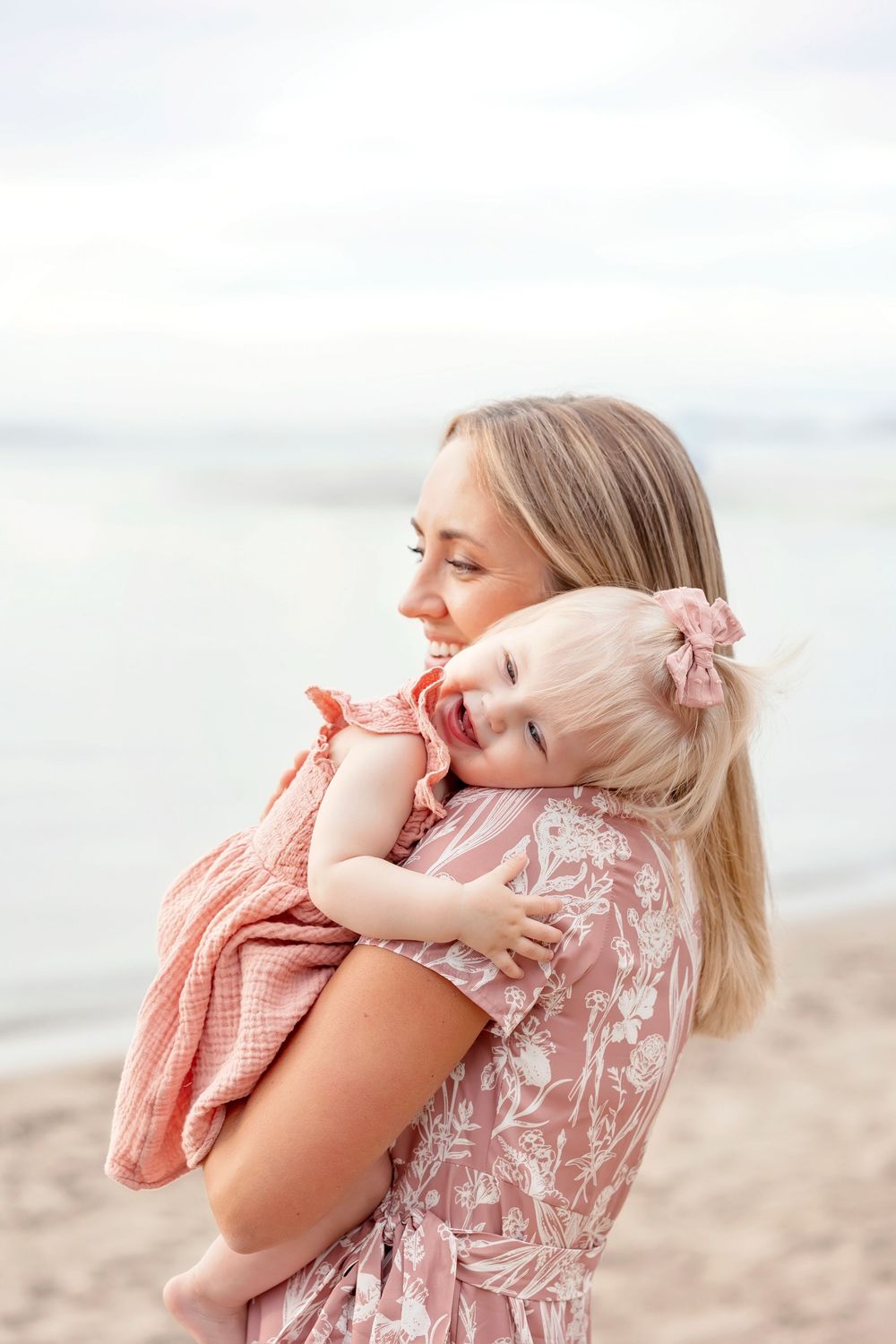 Mother and baby share tender moments on a sandy beach in matching pink floral outfits.