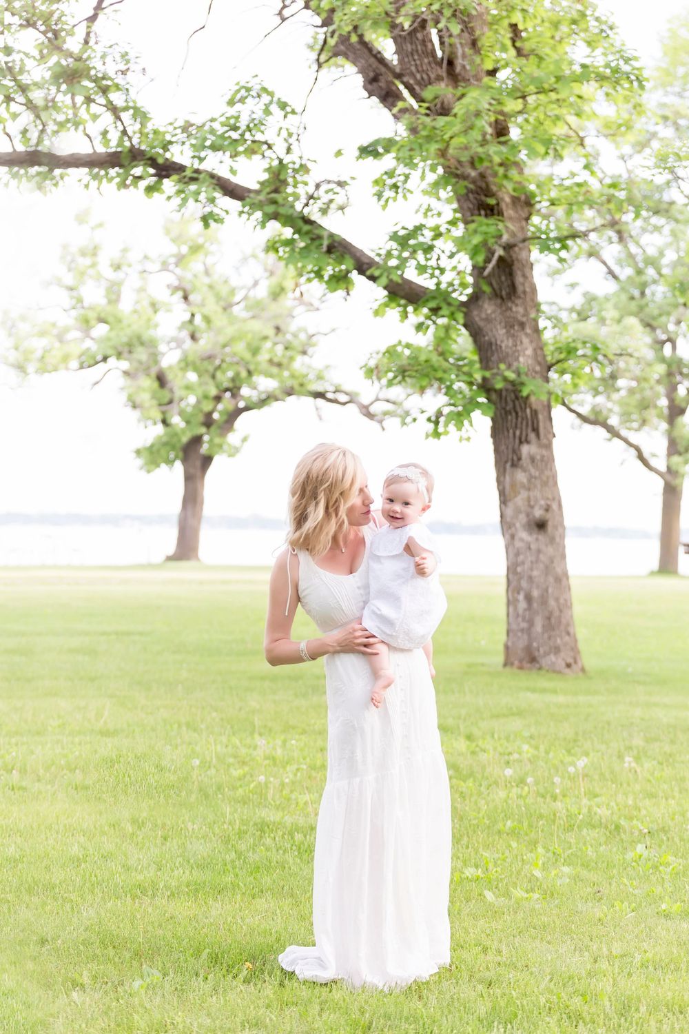 Parent in white dress holding laughing baby outdoors under green trees in sunlit park.