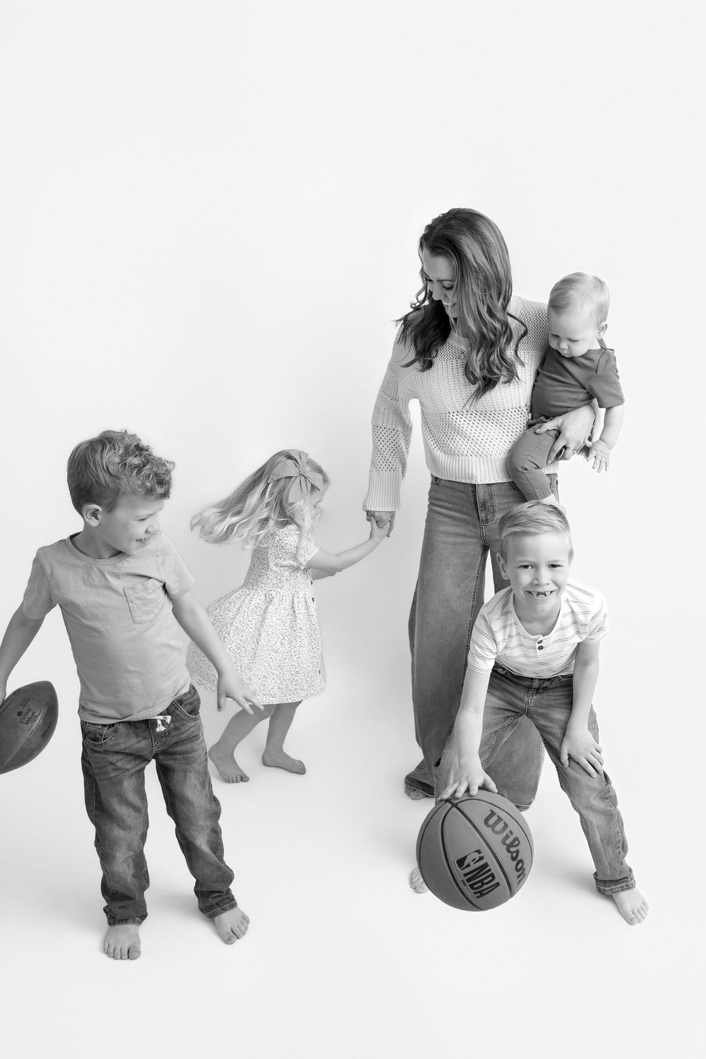 Black and white family portrait of a mother holding a baby with three children playing with a basketball and hat.
