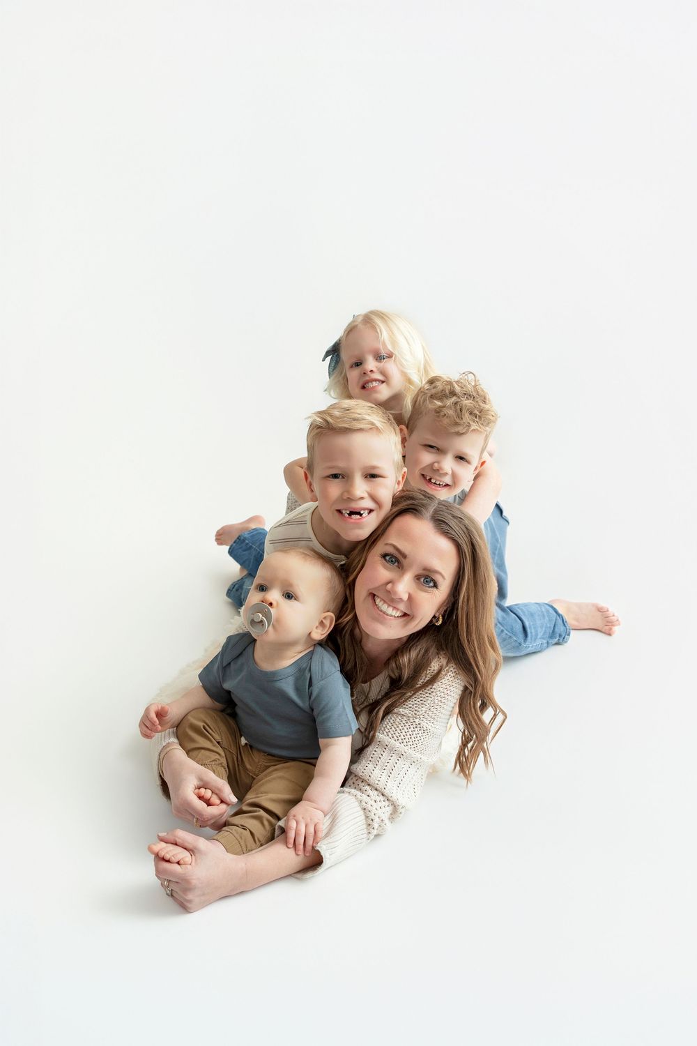 A smiling mother lies on a white floor with her four young children stacked and cuddled around her in a fun family portrait.