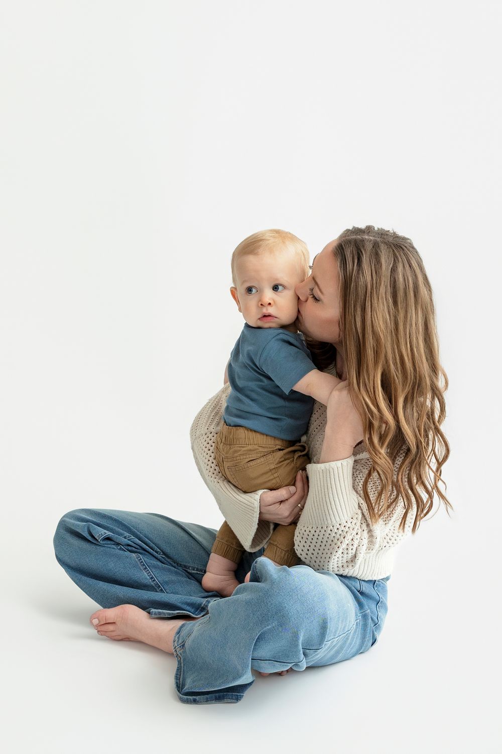 A young mother with wavy hair sits cross-legged kissing her baby boy, both wearing casual clothes on white background.