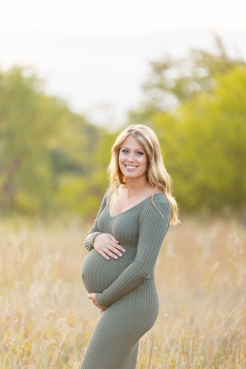 A woman in a green sweater poses in a golden autumn field during a maternity photoshoot.