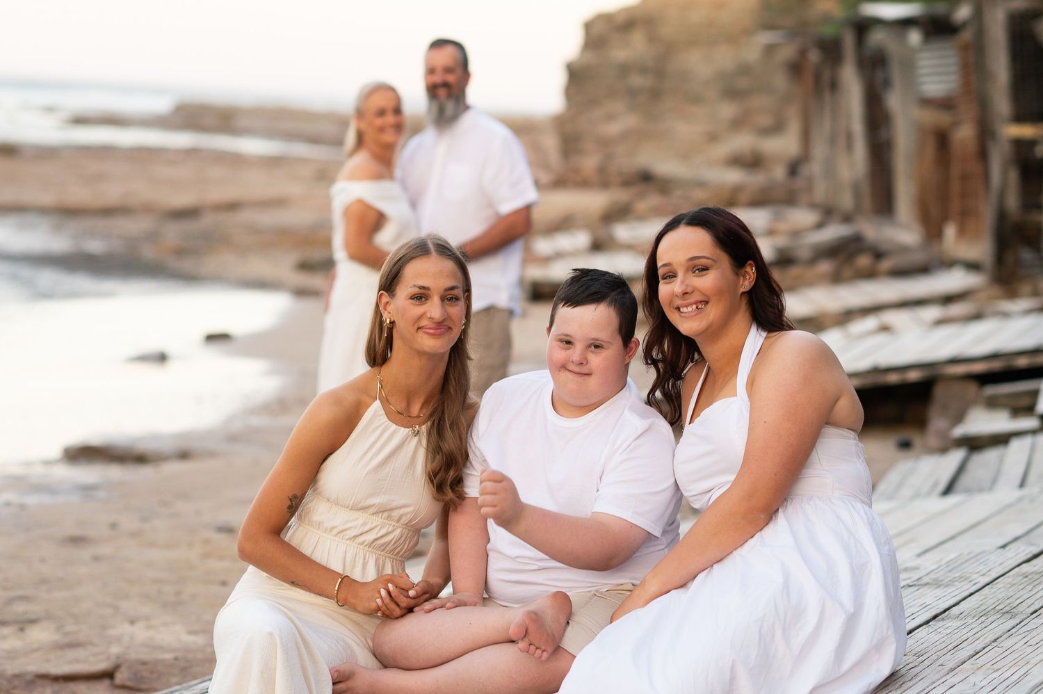 A family group dressed in white sits and stands on a rocky beach at sunset with cliffs in the background.