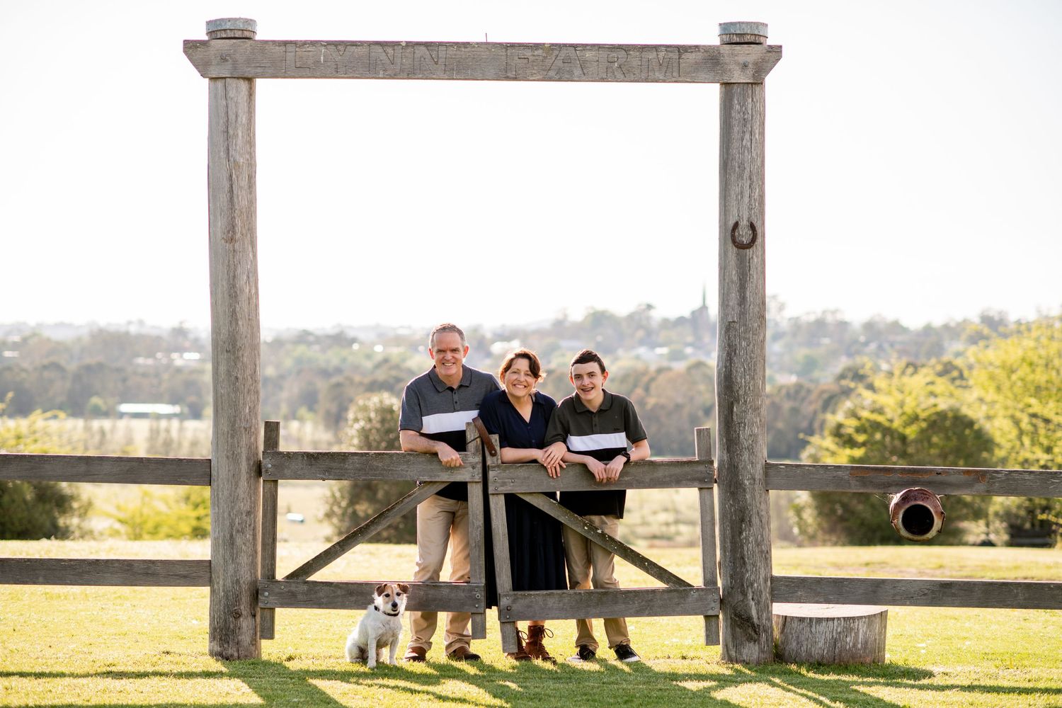 Three people stand together in front of a rustic wooden frame structure overlooking a scenic hillside landscape on a sunny day.