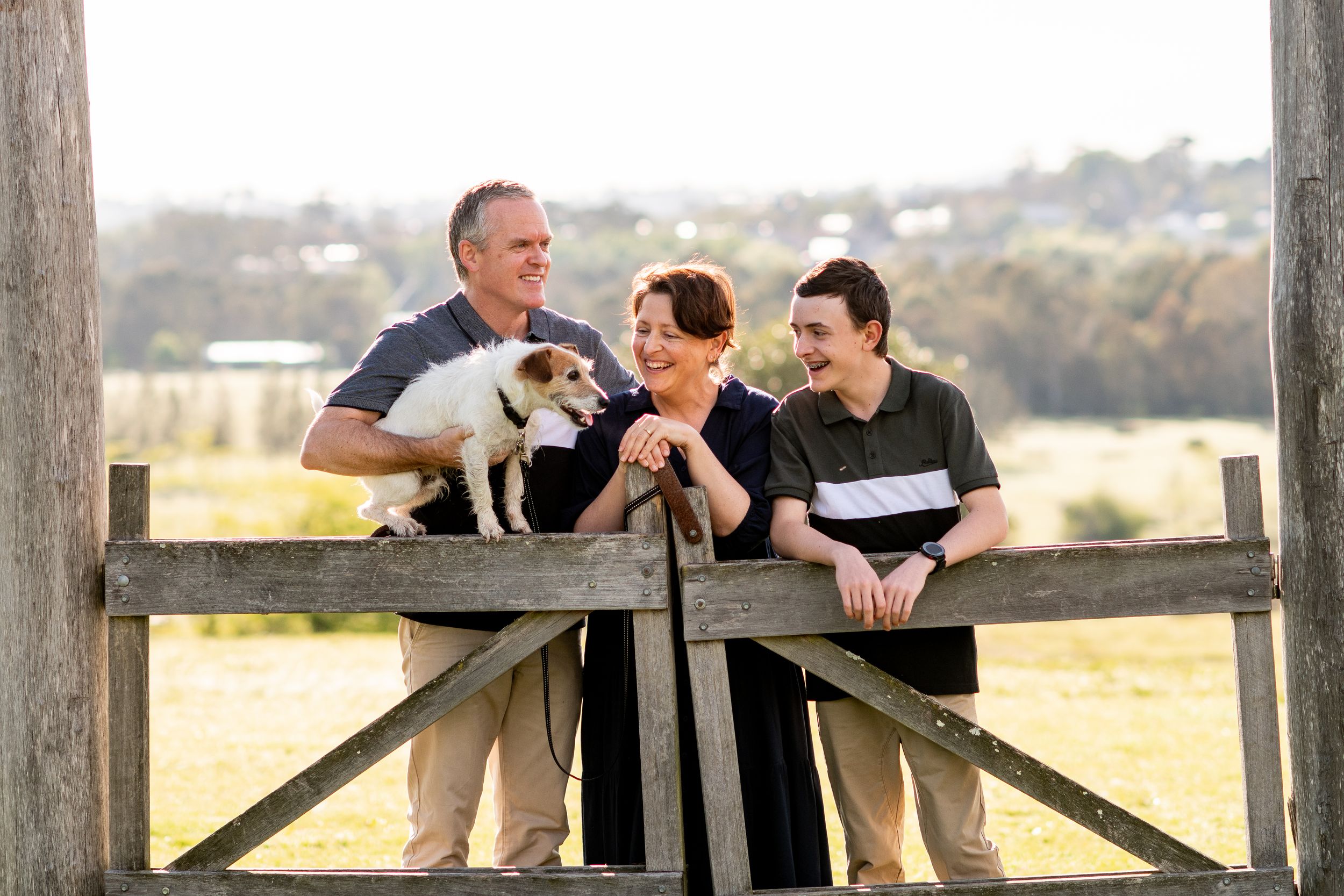 Three people stand at a wooden railing outdoors, sharing a joyful moment with a small white puppy.