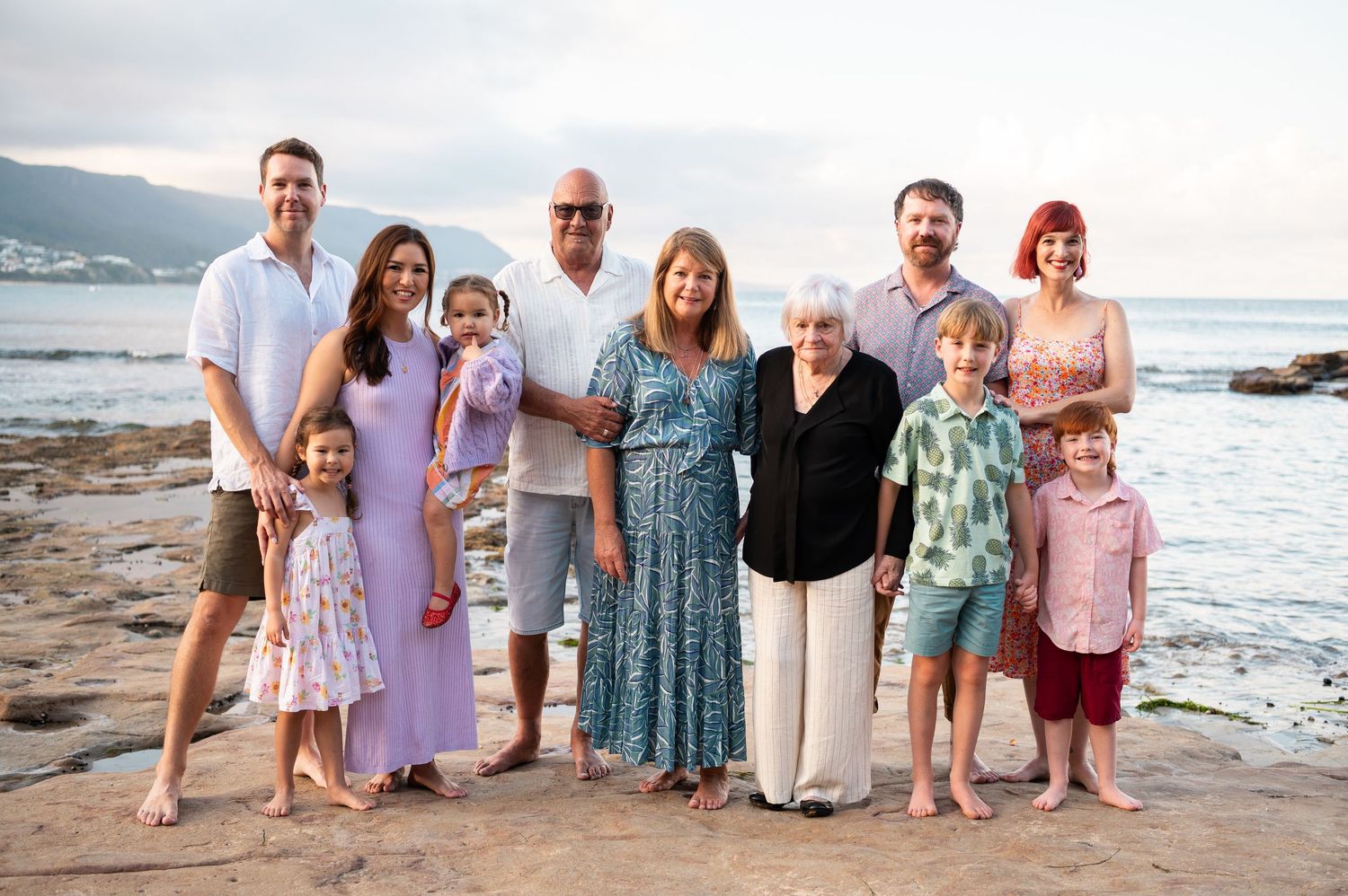 Multi-generational family group poses together on a rocky beach at sunset with mountains and ocean in the background.