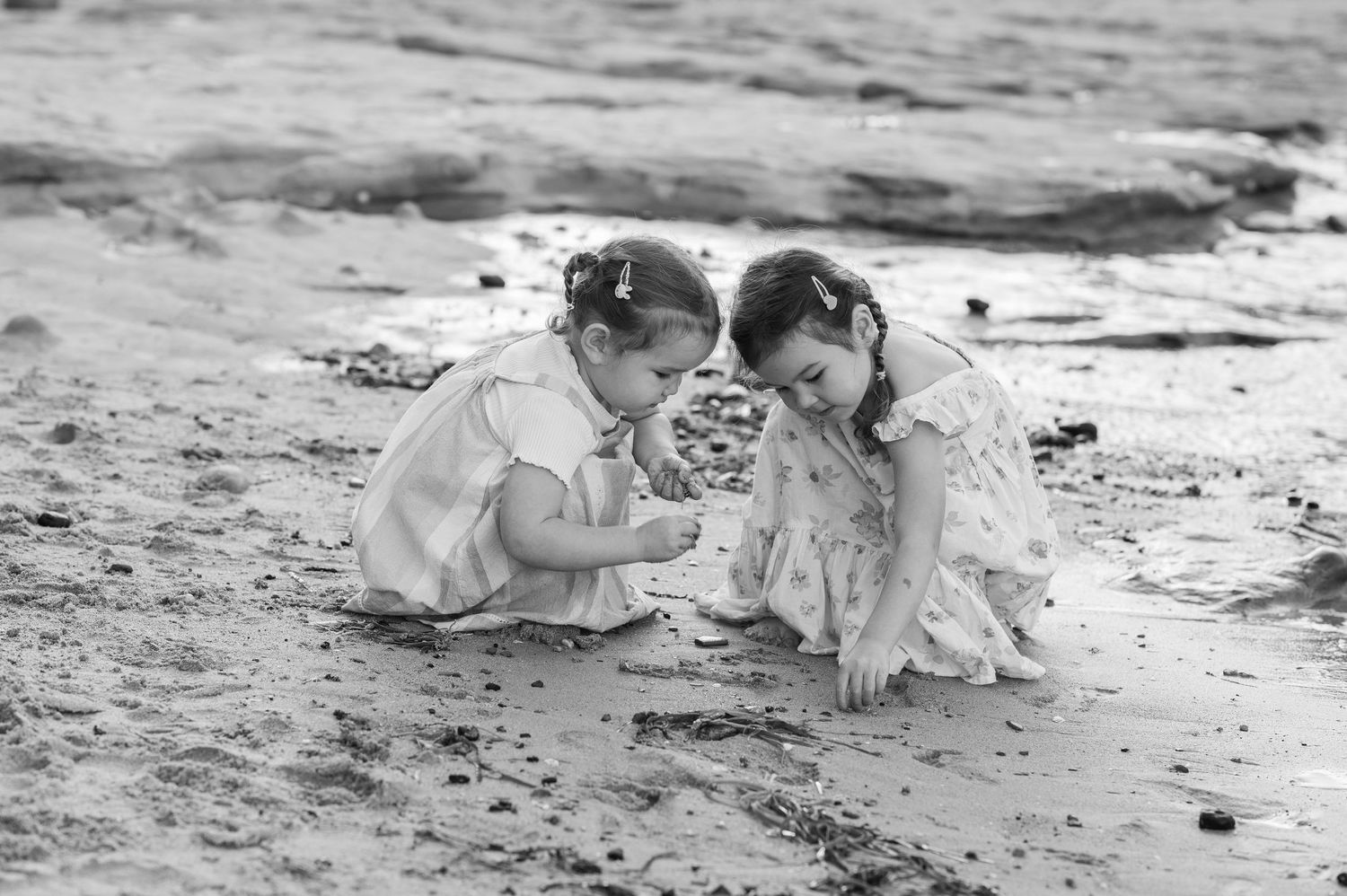 Two young children in white dresses crouch together at the beach, exploring something in the sand in black and white.