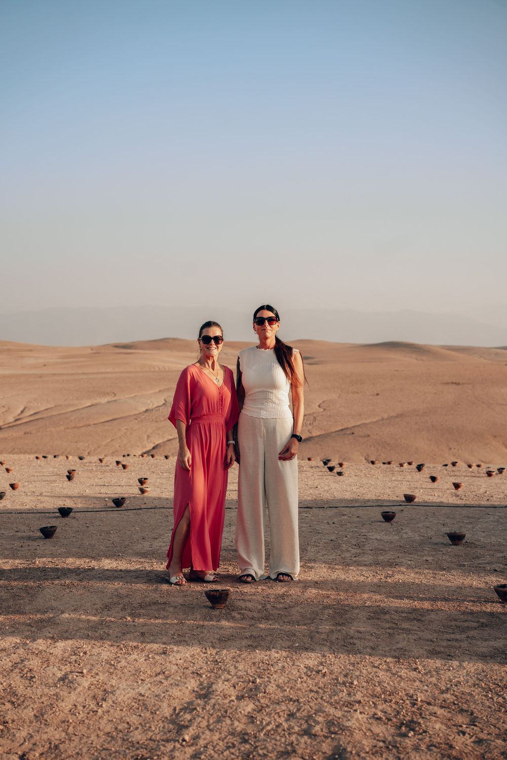 Two people in traditional desert attire stand together against a backdrop of sandy dunes in the Arabian desert.