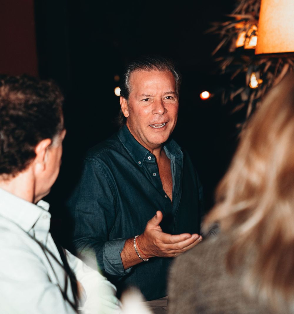 A person in a dark jacket laughs and gestures while engaged in conversation at a dimly lit social gathering.