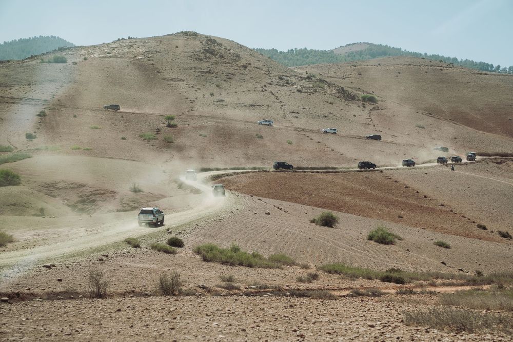Vehicles travel along a dusty dirt road winding through barren brown hills in a remote mountainous landscape.