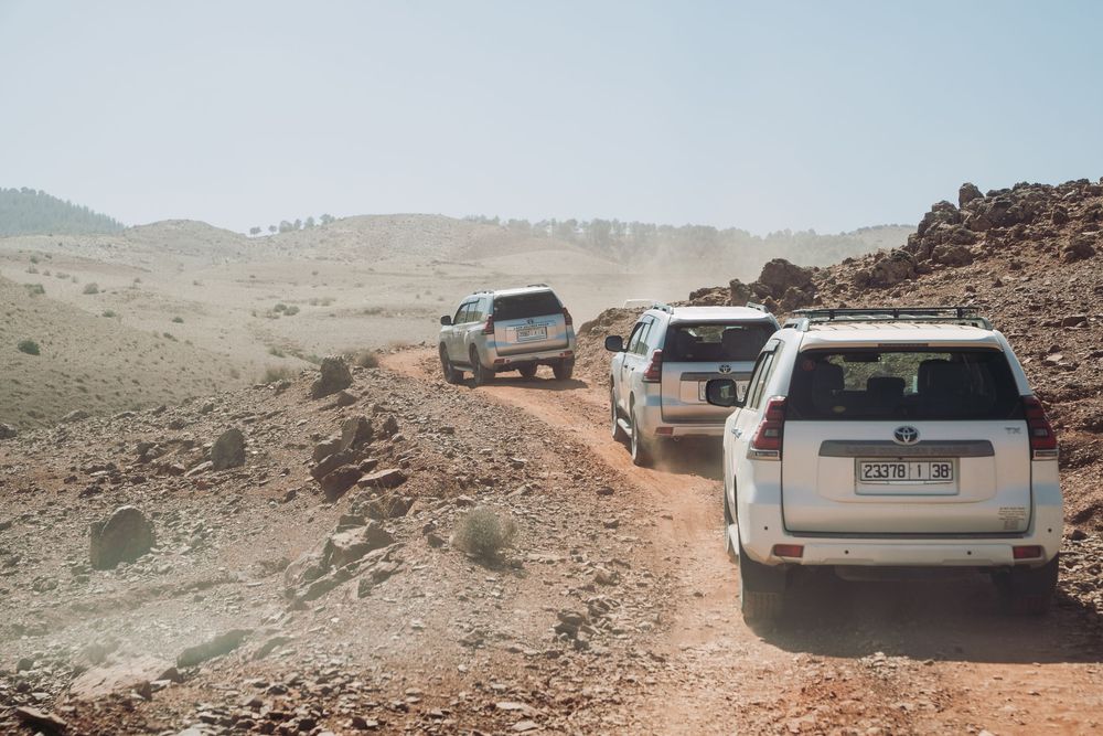 Three white SUVs driving in a line on a dusty mountain trail through rugged desert terrain.