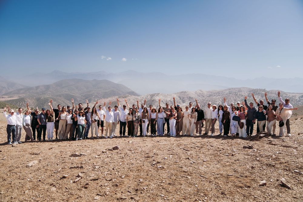 A large group of people stand together with raised arms on a desert mountain ridge against a hazy landscape backdrop.