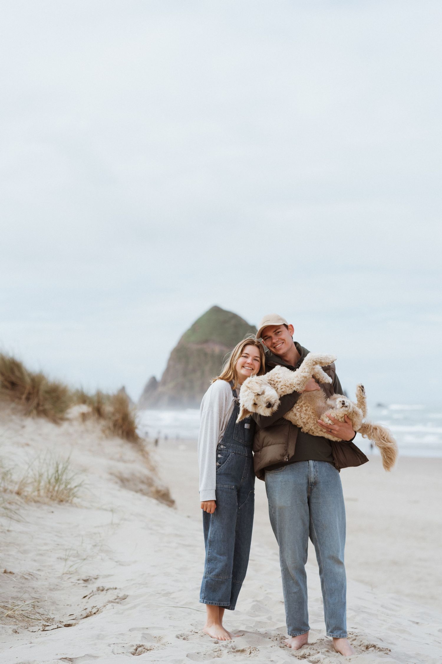 Couple holding dogs while standing on beach with Haystack Rock visible in background at Cannon Beach.