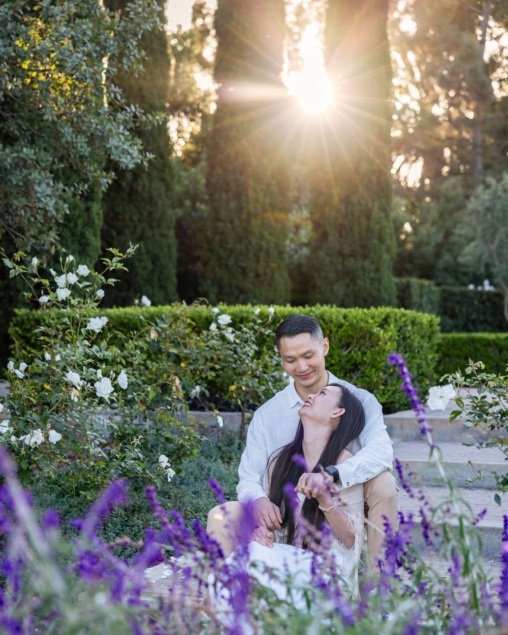 at the Laguna Niguel botanic romantic couple sits together in a sunlit garden surrounded by purple lavender and white roses at golden hour.