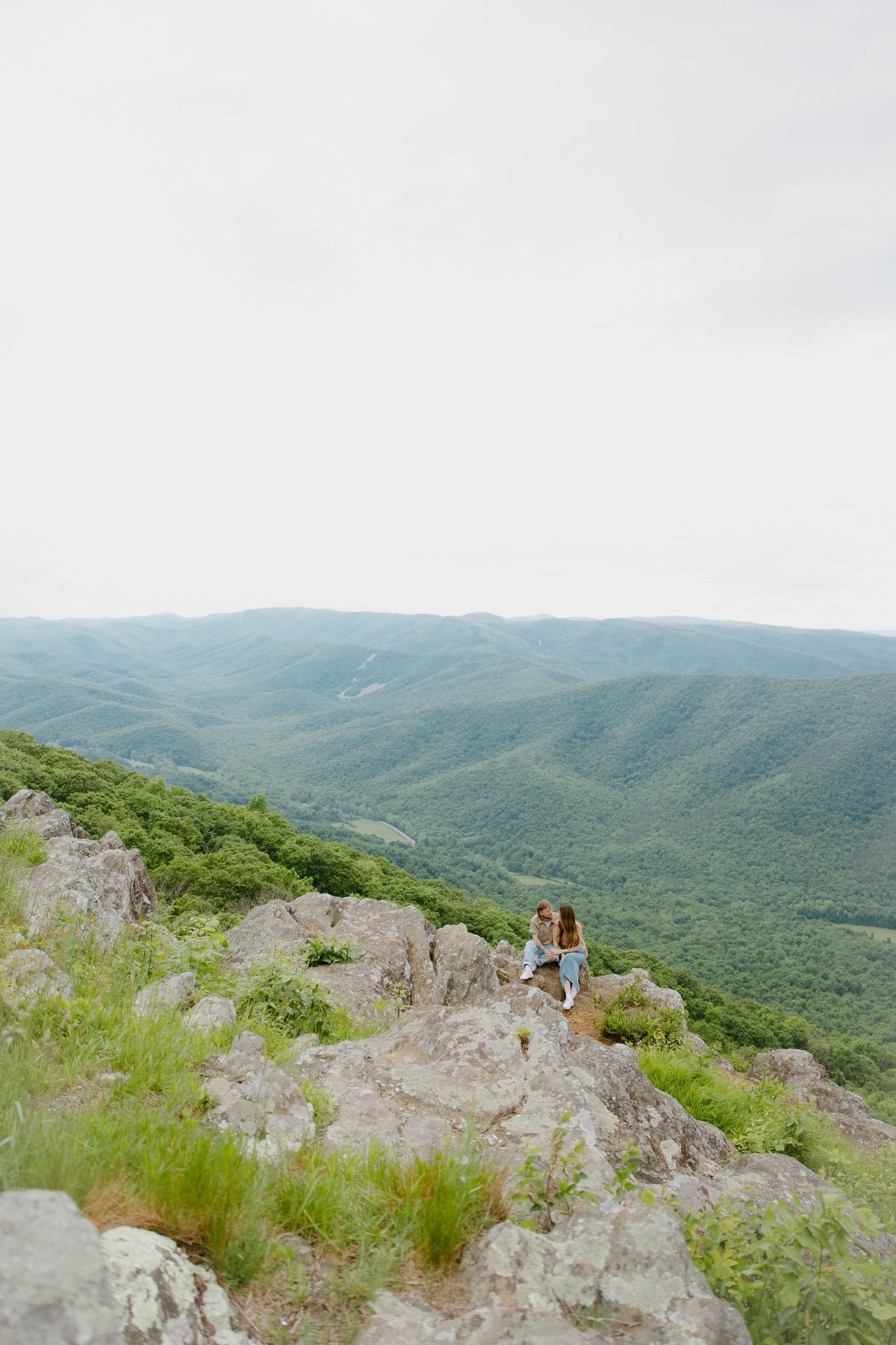 Two people standing on rocky mountain overlook with vast green valley views.