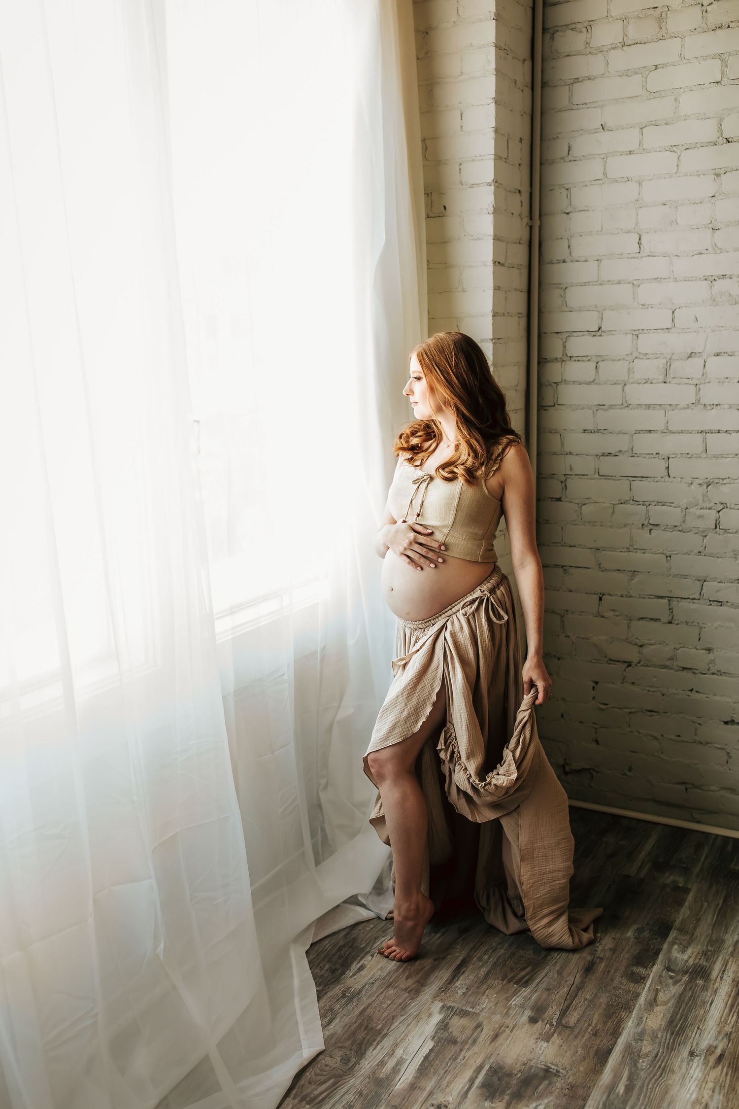 An expectant mother in a flowy dress stands by a sunlit window against a white brick wall in an artistic maternity photo.