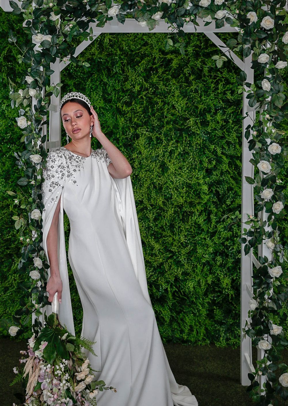 Bride in white cape gown with crystal embellishments poses under floral arch with green ivy wall backdrop.