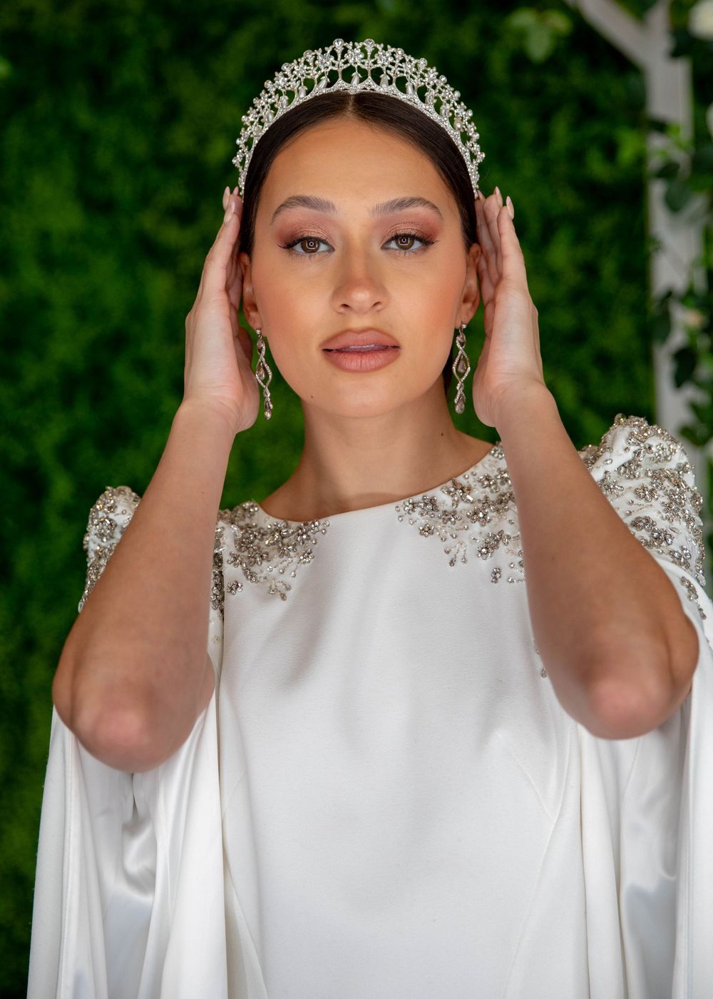 Bride wearing a crystal tiara and white gown with beaded shoulders, posing with hands near her head.