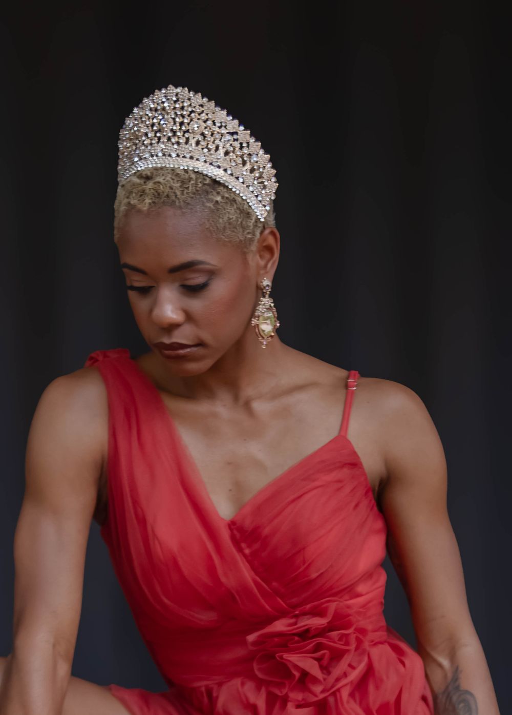 Woman in red dress wearing crystal crown with short blonde hair, looking down elegantly.