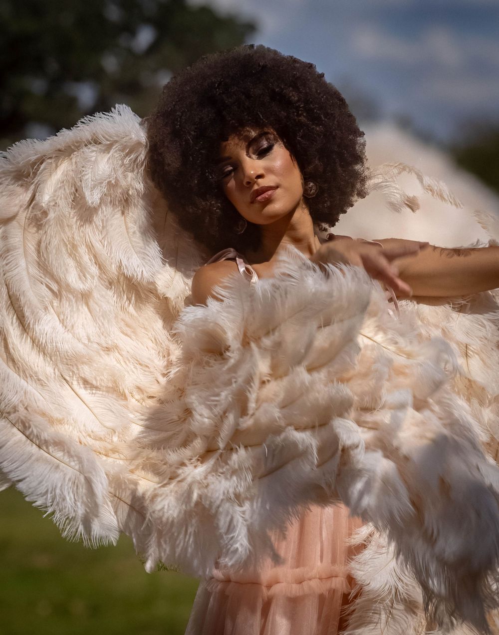 A woman with a curly afro poses outdoors wearing a stunning feathered cape in cream and blush tones.