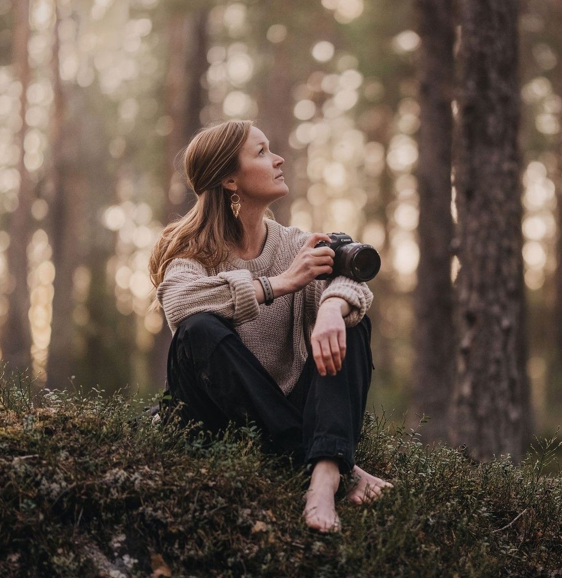 Young woman photographer sitting barefoot on mossy forest floor holding camera looking up.