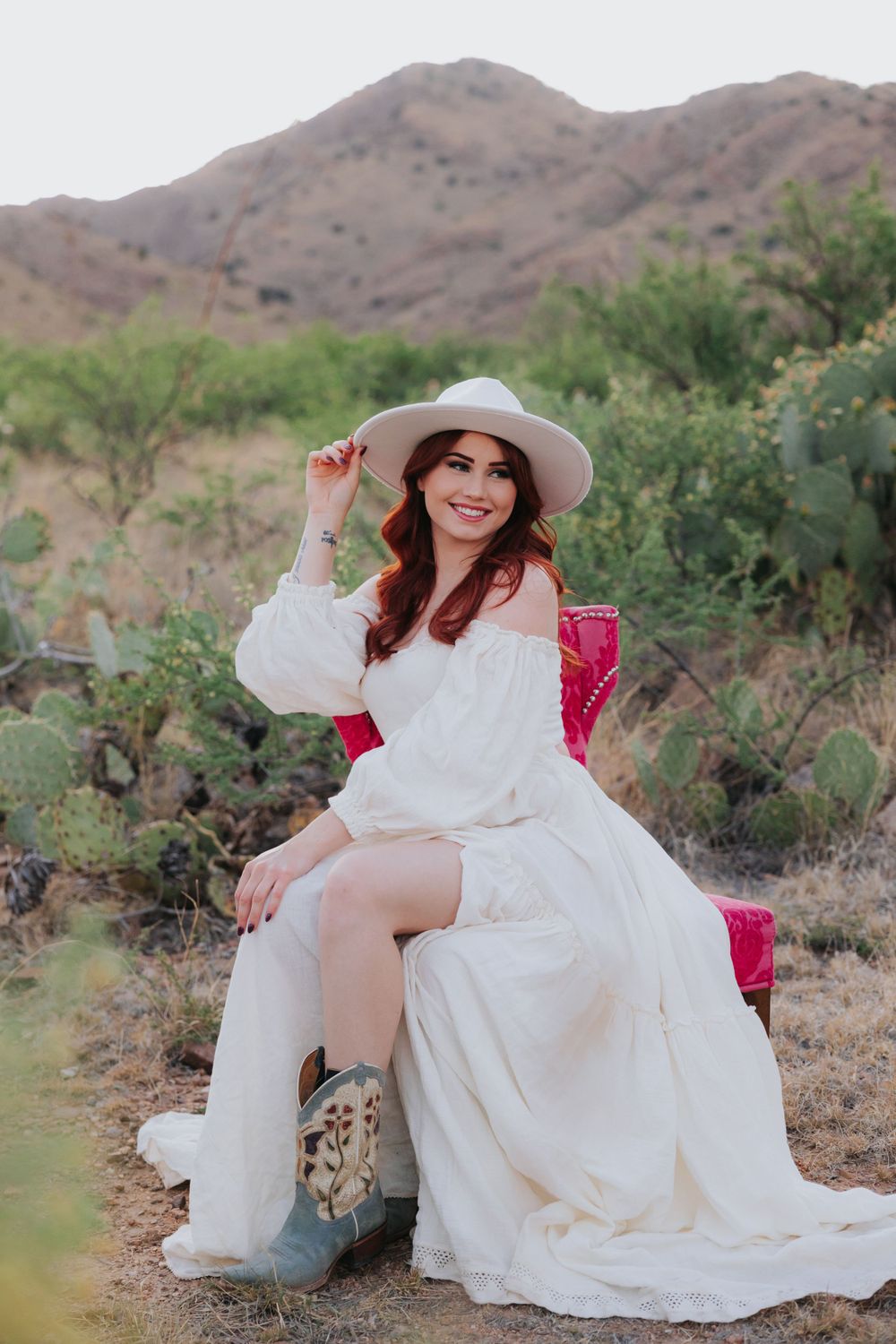 Woman in white dress posing on pink chair in desert setting.