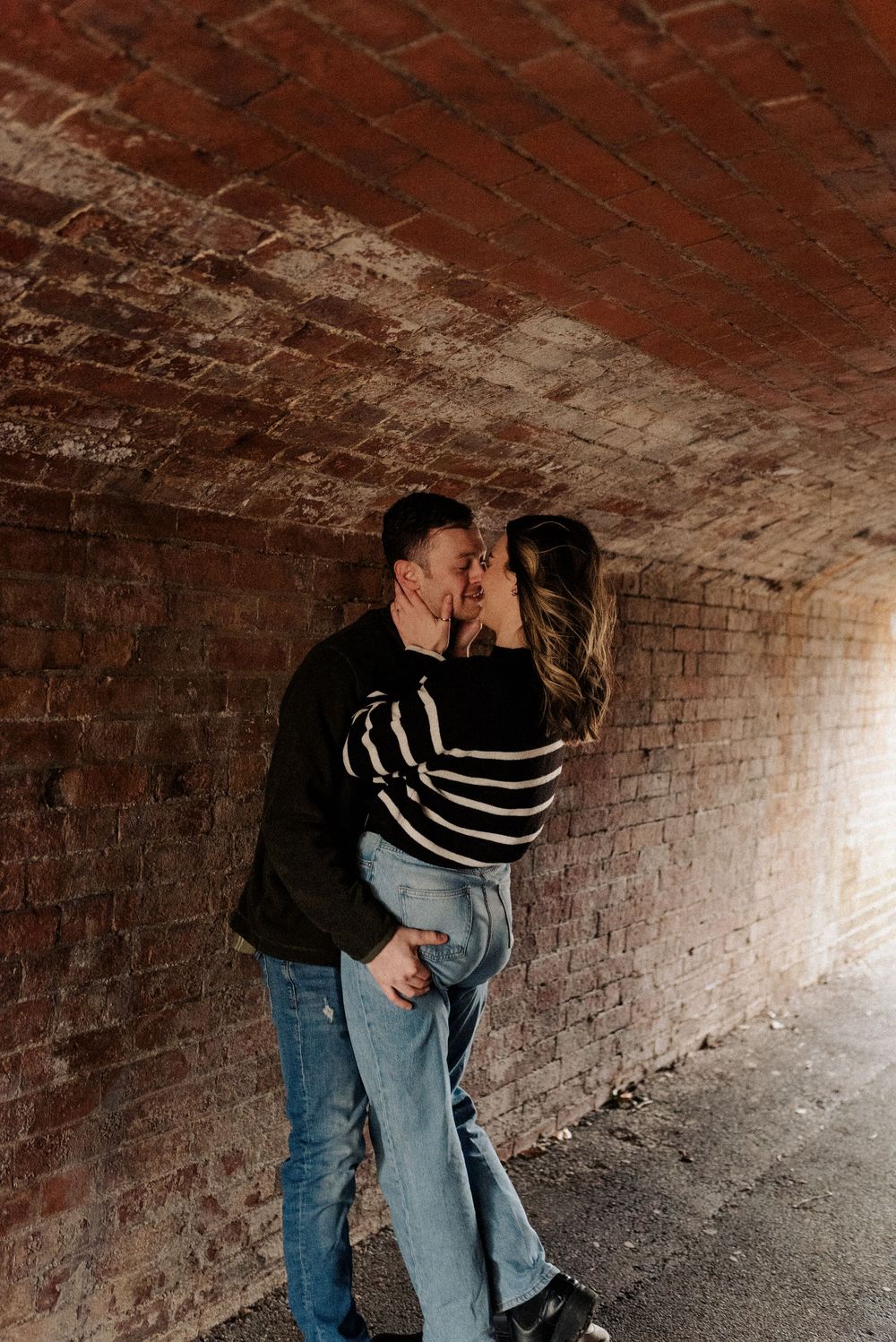A couple embraces and kisses in a dimly lit brick tunnel.
