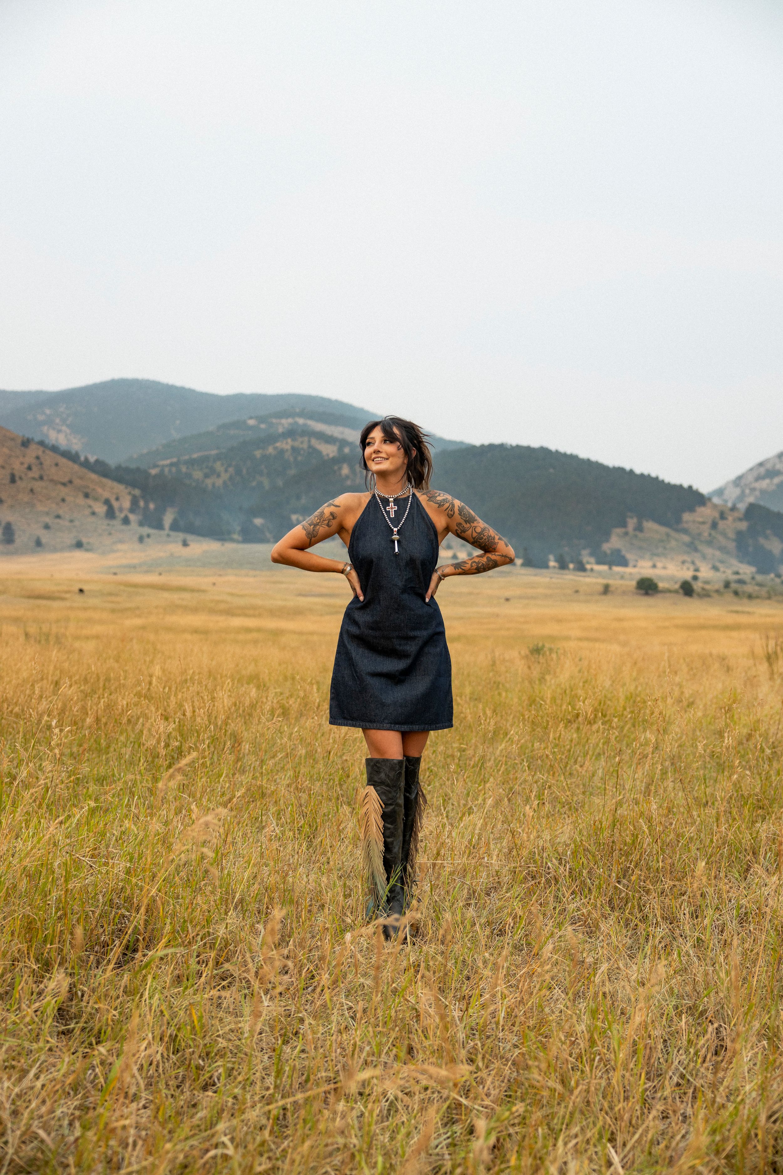 Tattooed woman in black denim dress and knee-high boots standing in golden field with mountain backdrop.