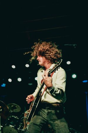 Energetic musician with curly hair playing guitar on stage with dramatic lighting.